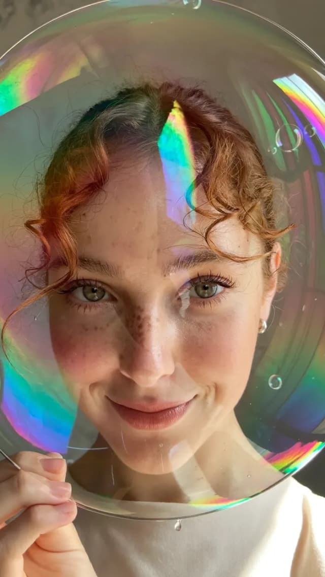 Femme rousse bouclée aux taches de rousseur souriant à travers un grand film de savon irisé – Higgsfield Soul 2